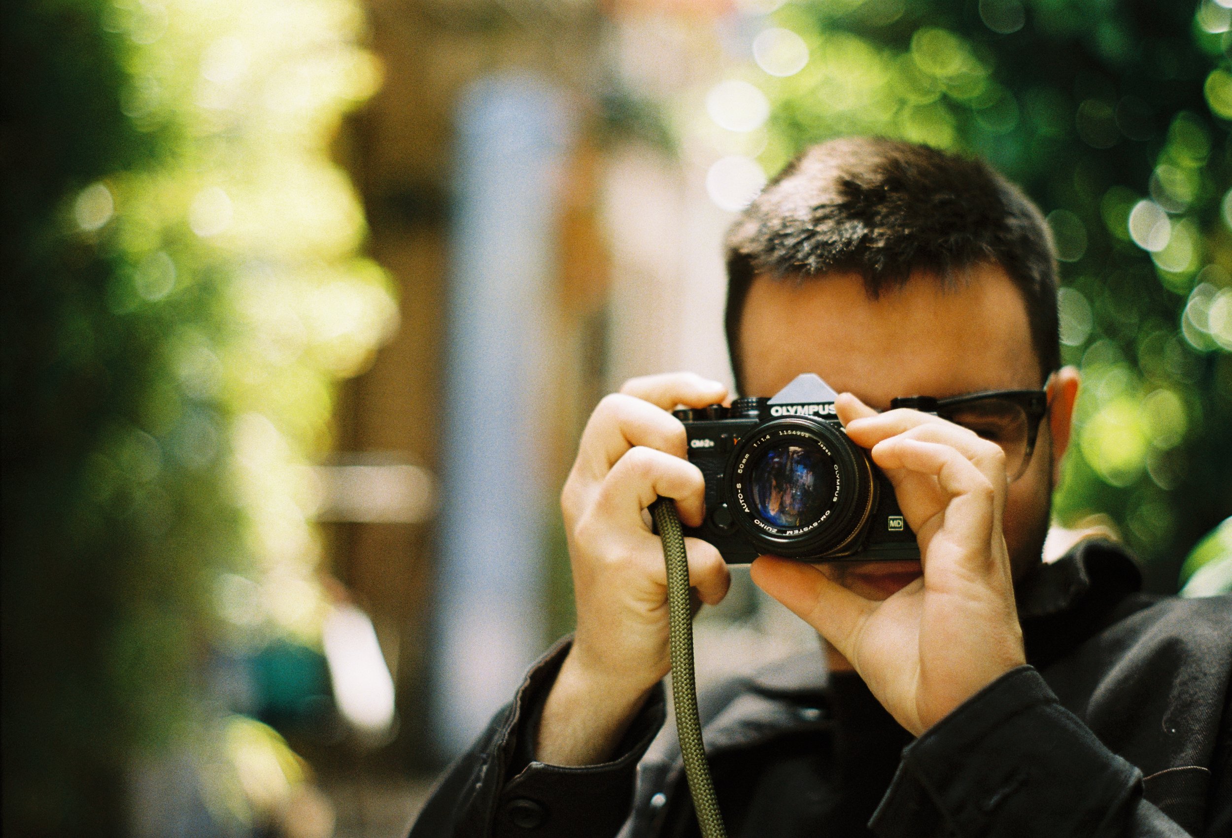 A person with short dark hair and glasses is taking a photo with a digital camera outdoors, with leafy greenery and sunlight in the background.