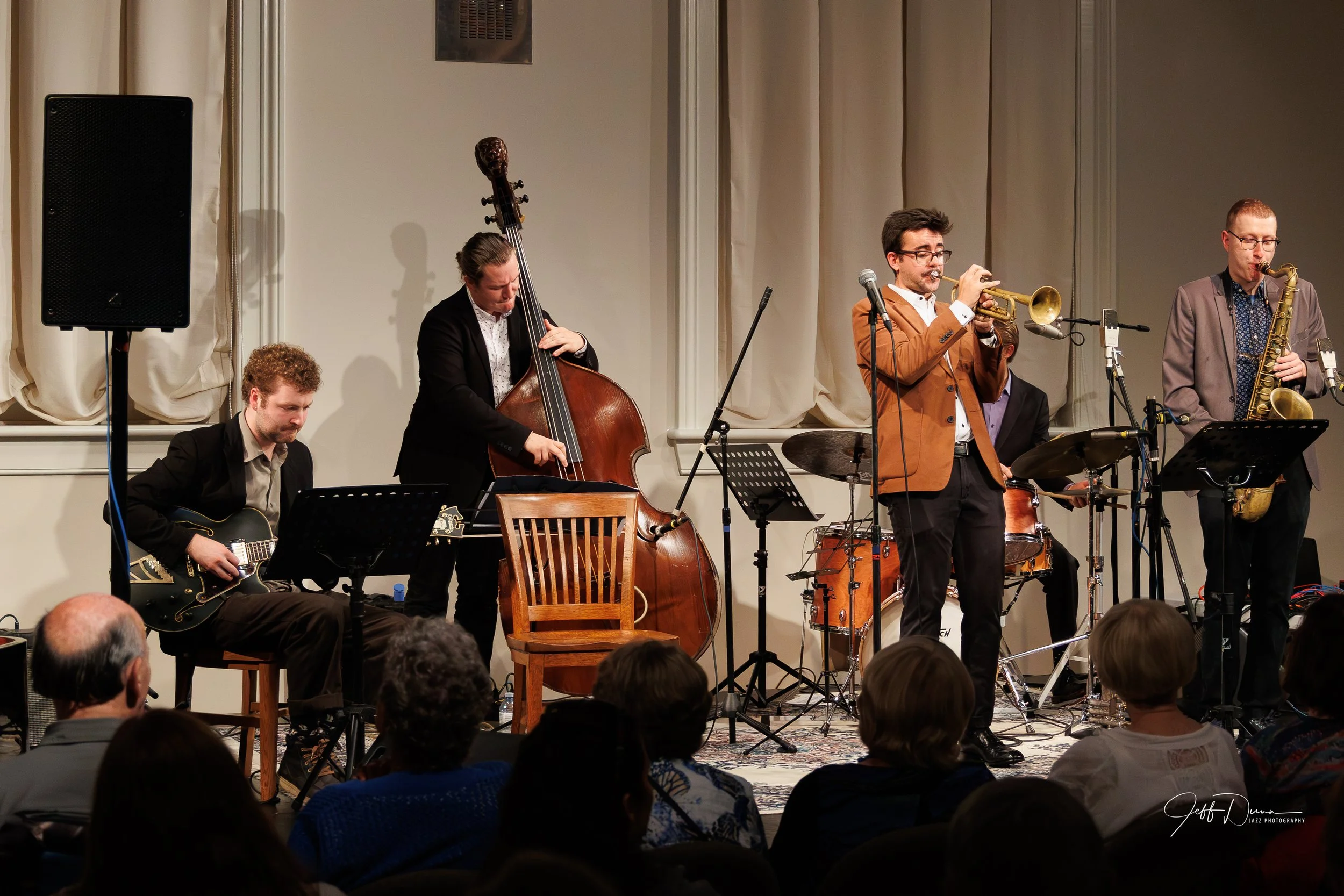 Jazz band performing on stage with five musicians playing guitar, double bass, trumpet, and saxophone, while audience watches.