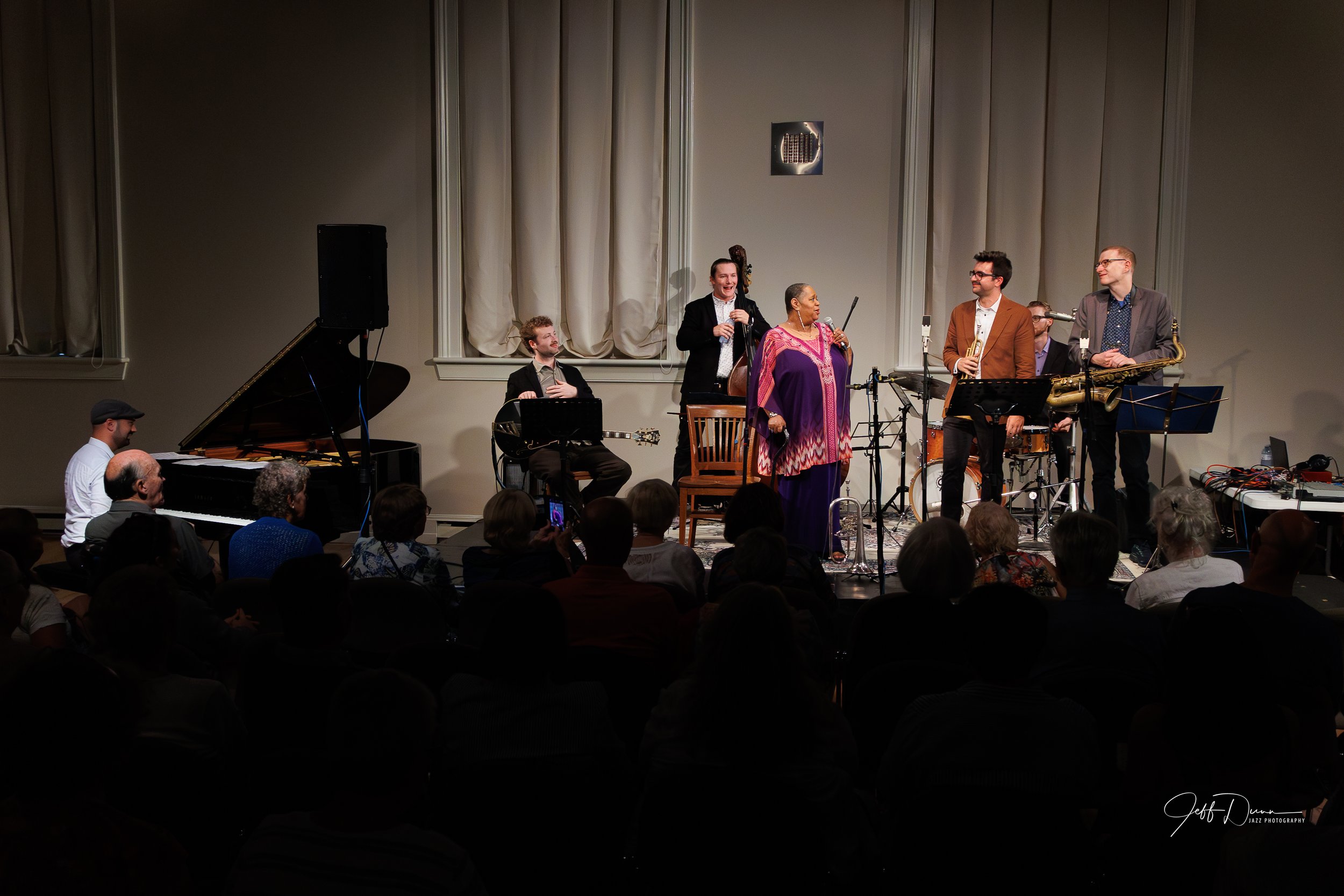 A jazz band performs on stage with a pianist, saxophonists, a percussionist, and a vocalist dressed in purple, in front of an audience.
