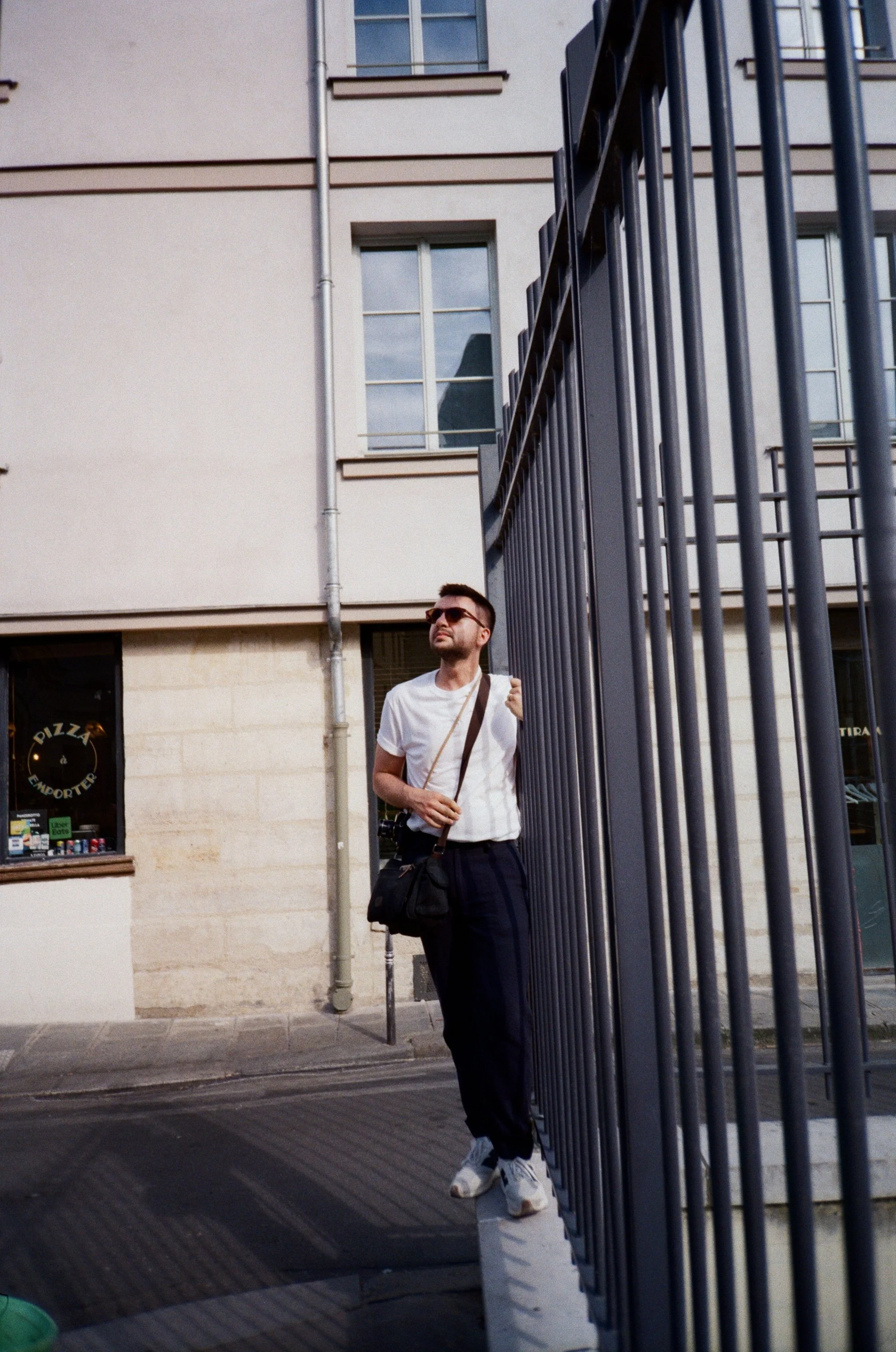 A man standing outside on the sidewalk near a black metal fence, wearing sunglasses, a white shirt, black pants, and sneakers, with a bag across his shoulder, looking to his left.