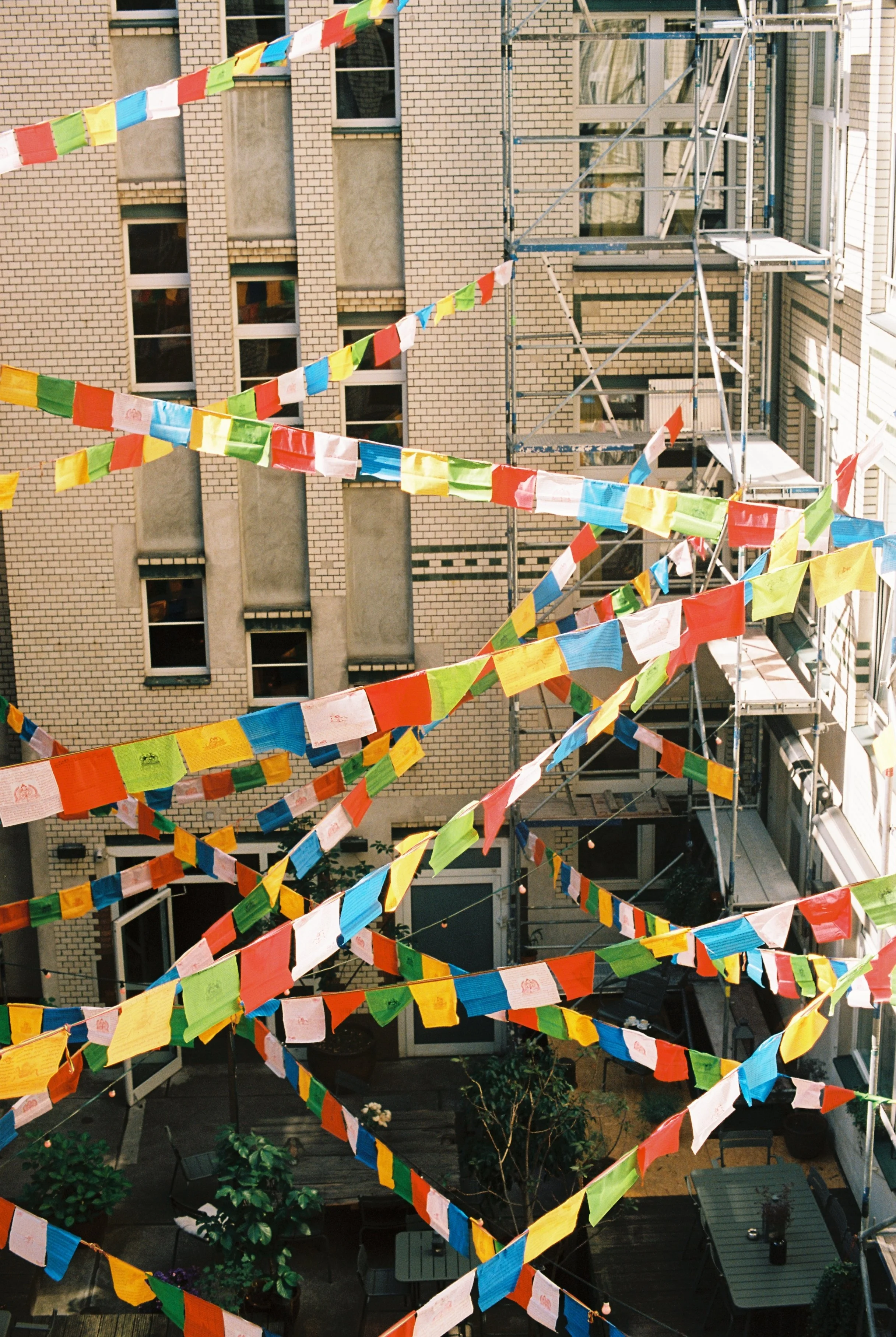 Colorful flags hanging across an outdoor courtyard surrounded by brick buildings, with scaffolding visible on one building and patio furniture below.