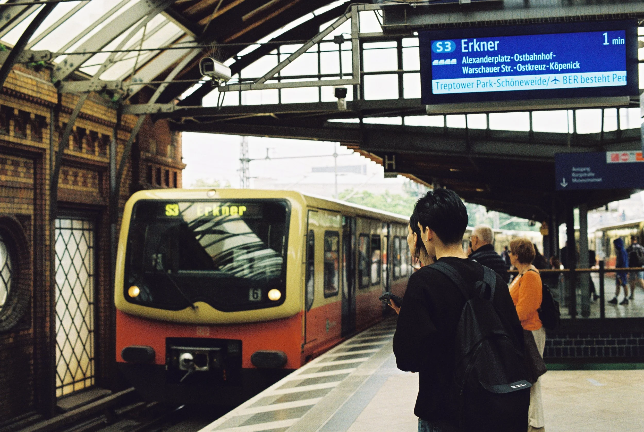 People wait for the S3 train at a station platform with a yellow and orange train stopped nearby, overhead digital display showing train information, some passengers standing, some using phones, and the station structure in the background.