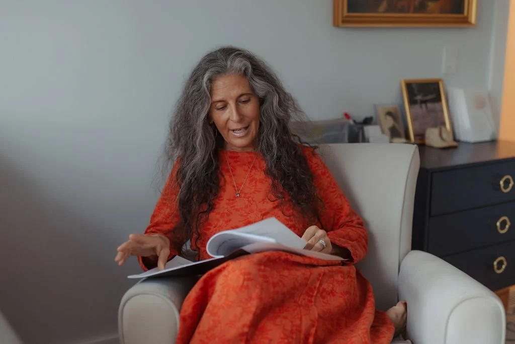 A woman with long gray hair sitting in a white armchair, wearing a red dress, reading a notebook or planner.