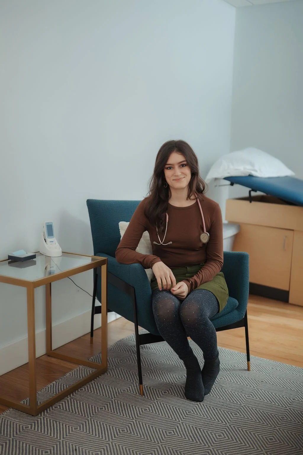 A young woman sitting in a blue chair in a medical office, with a stethoscope around her neck, smiling at the camera. There is a small table beside her with a phone and a device on it, and medical examination table in the background.