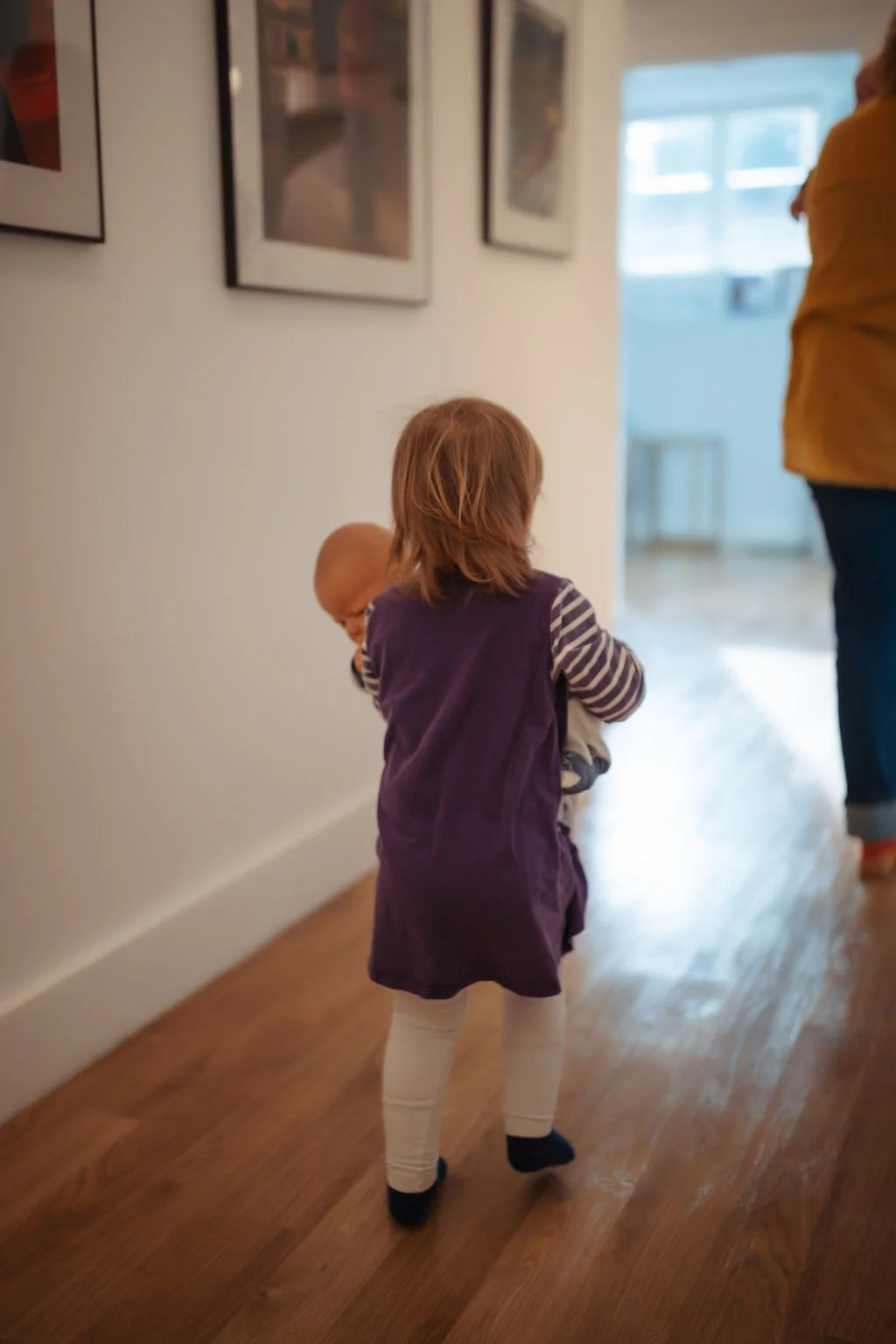 A young girl with brown hair, dressed in a purple dress, striped long-sleeve shirt, and white leggings, is holding a small baby baby while walking down a hallway with wooden floors inside a home. A person in a yellow top and blue jeans stands nearby,