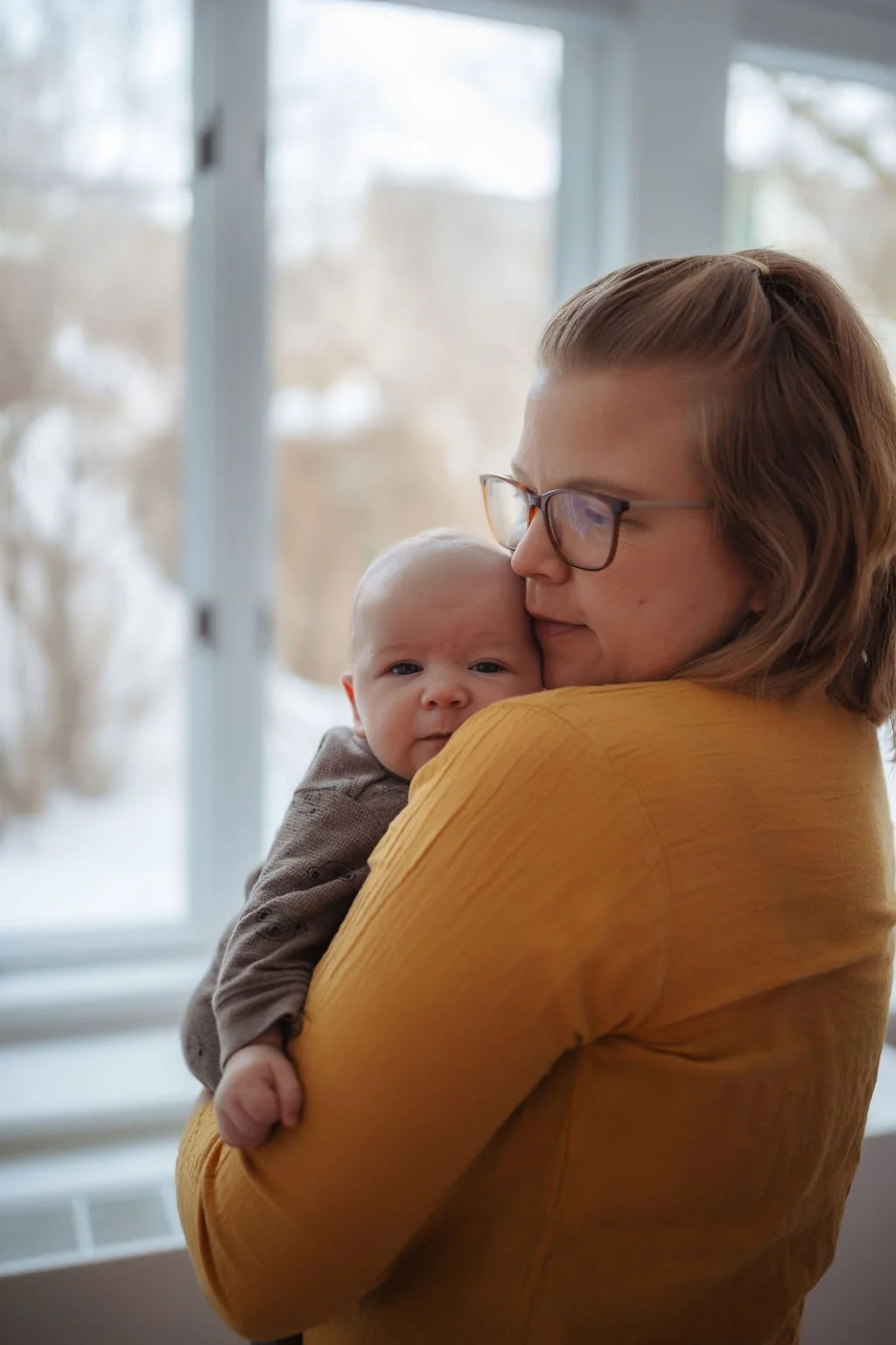 A woman with glasses and a yellow top holding a baby close to her face indoors, with a window in the background.