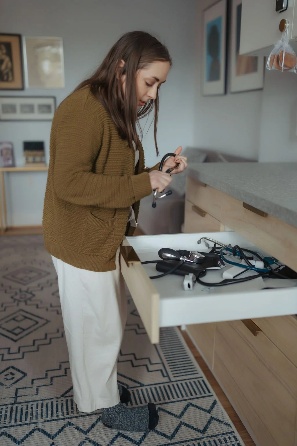 A woman dressed in a brown sweater and white pants looking into an open drawer filled with medical equipment, including a stethoscope, blood pressure cuff, and other medical devices, in a cozy home setting.