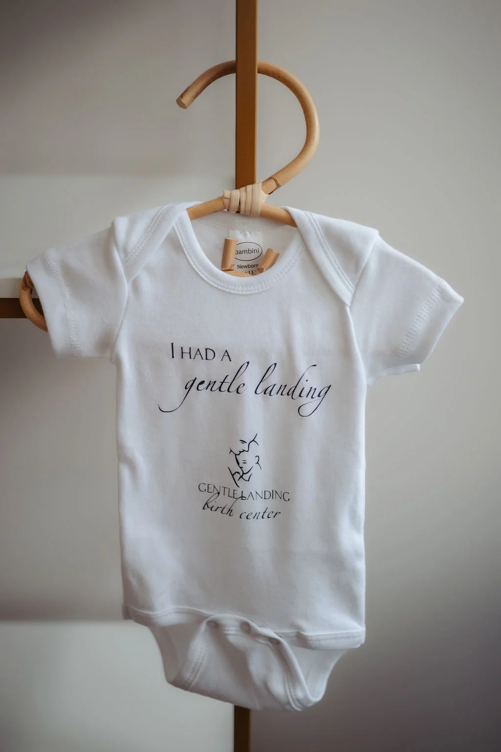 White baby onesie hanging on a wooden hanger with the text 'I had a gentle landing' and a small illustration of a person holding a baby, with additional text 'Gentle landing birth center'.
