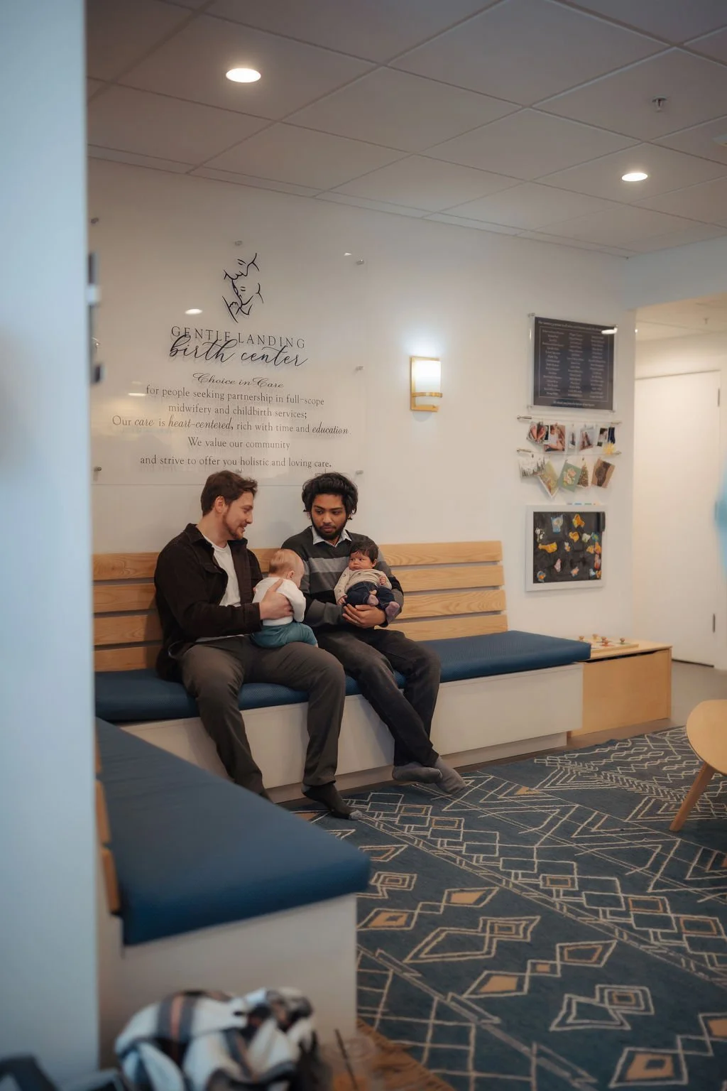Two men sitting on a wooden bench in a waiting area, each holding a baby. The background has a wall with decor, a wall sconce, and signage for a birth center.