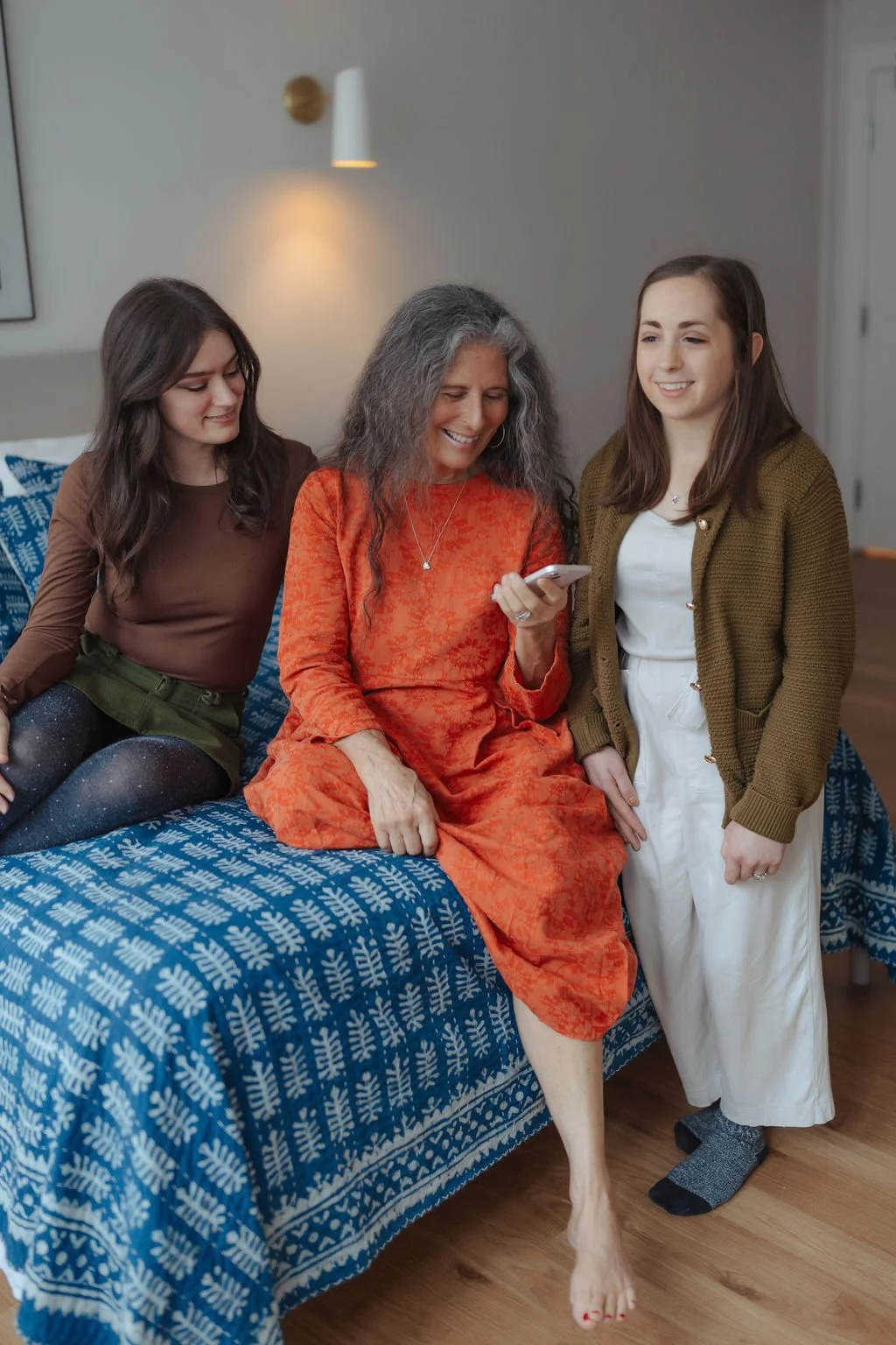 Three women sitting and standing on a patterned blue couch, smiling and looking at a smartphone.