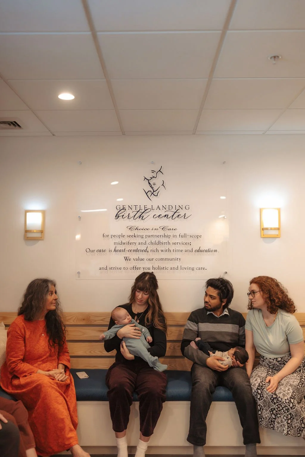 People sitting on a bench inside the GENTLE LANDING birth center, holding babies, with a sign on the wall behind them.