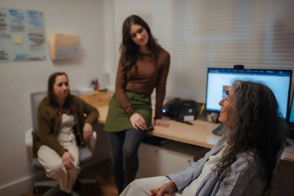 Three women in a medical office, one older woman sitting and two younger women standing and sitting nearby, with computer monitors on the desk.