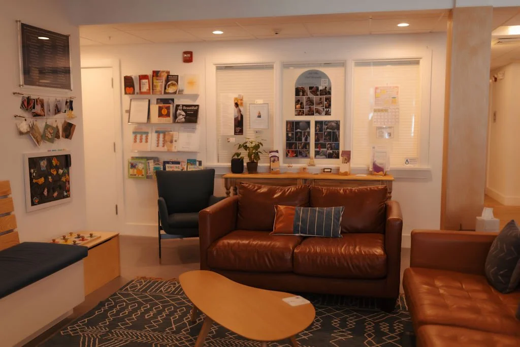 A cozy waiting room with brown leather couches, a black armchair, a small wooden table, and wall shelves with magazines and decorations. There is a window with blinds and a bulletin board with photos and papers on the wall.