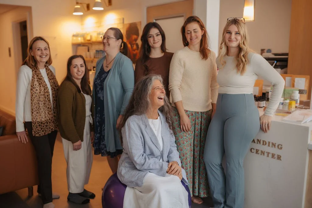 Seven women, including an elderly woman sitting on a purple exercise ball, are gathered in a warmly lit room, smiling and laughing together.