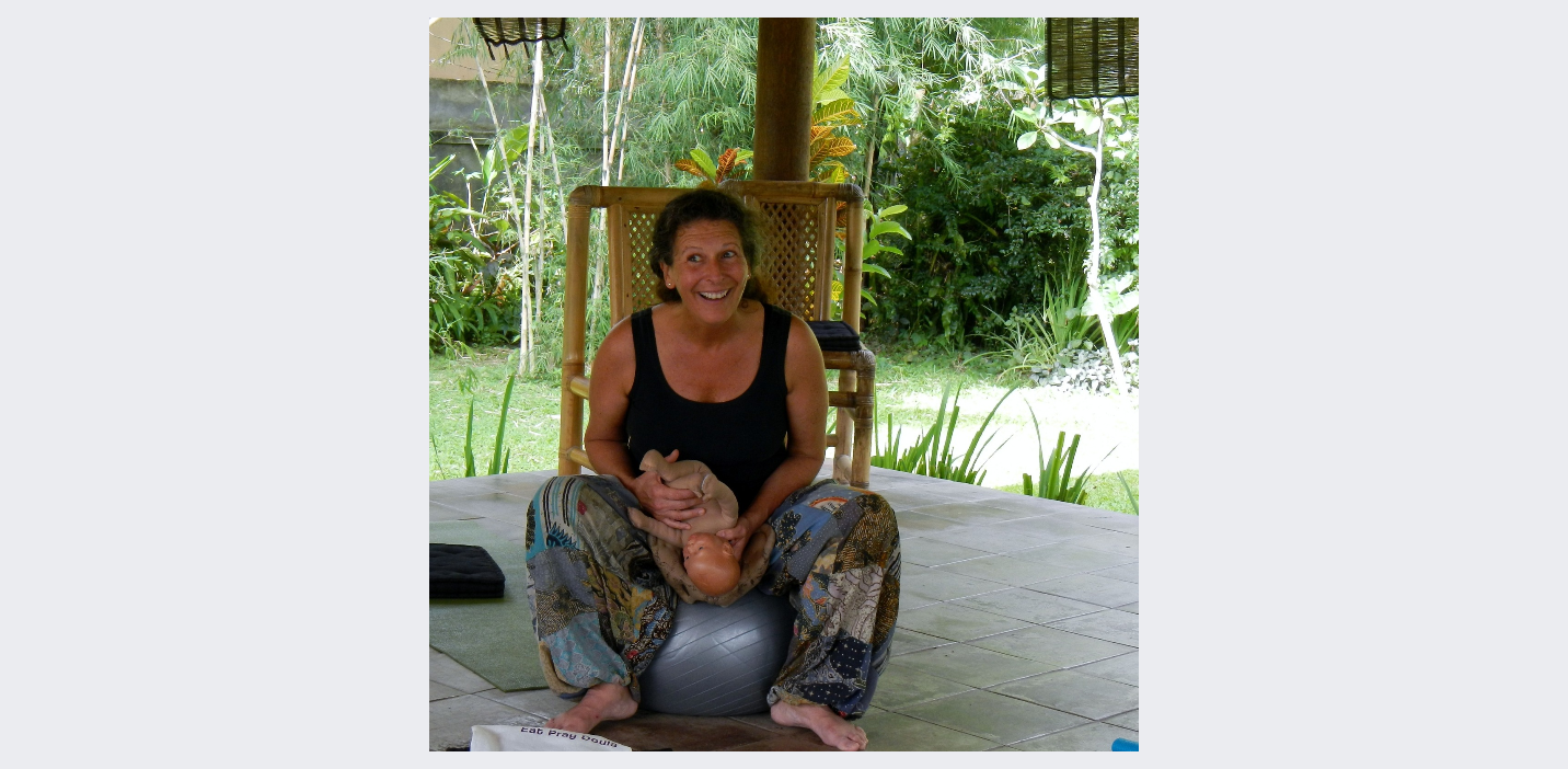 A woman sitting on an exercise ball, holding a baby doll, and smiling outdoors on a covered porch with green foliage in the background.