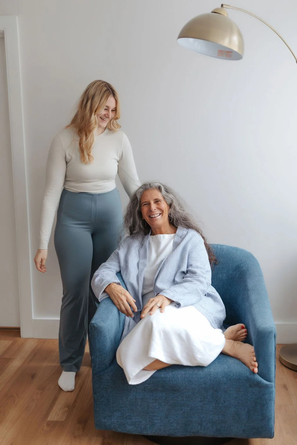 A woman with long gray hair sitting comfortably in a blue armchair, smiling, wearing a white dress and a light cardigan, with a younger woman with blonde hair standing beside her, smiling and gently touching her shoulder, in a bright room with white 