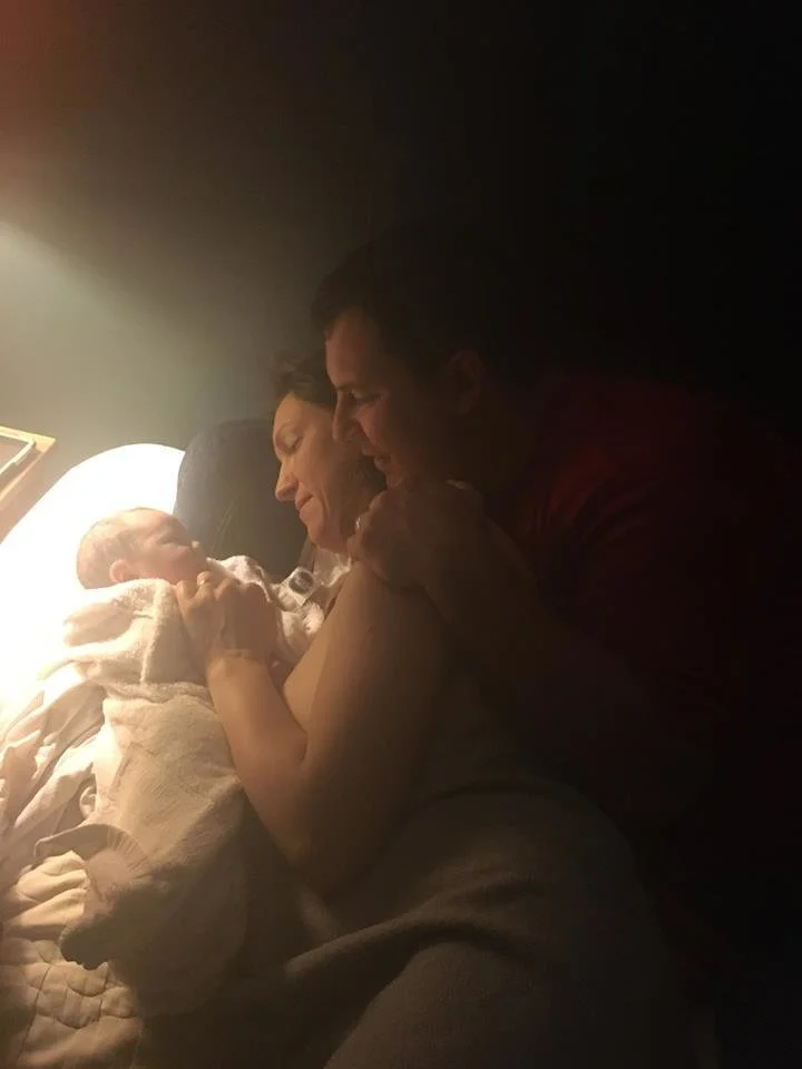 A family in a hospital room with a newborn baby. The mother is lying in bed holding the baby, and the father is leaning close, smiling and touching the mother and baby.