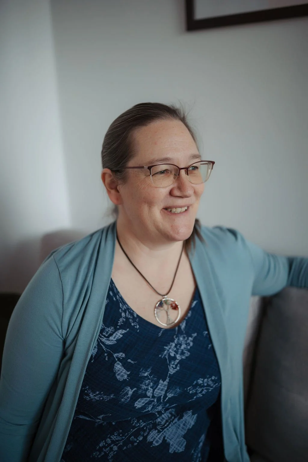 A smiling woman with glasses, wearing a blue patterned top and a necklace, sitting on a couch in a living room.