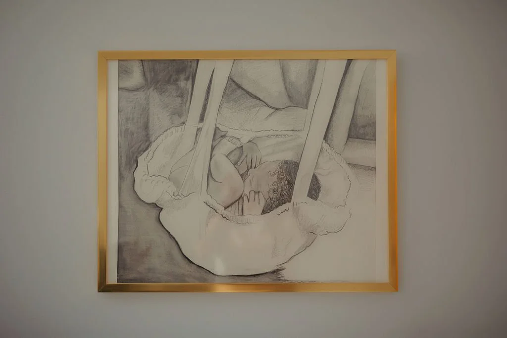 Drawing of a newborn baby in a hospital bassinet with a blanket, on a wall framed in gold.