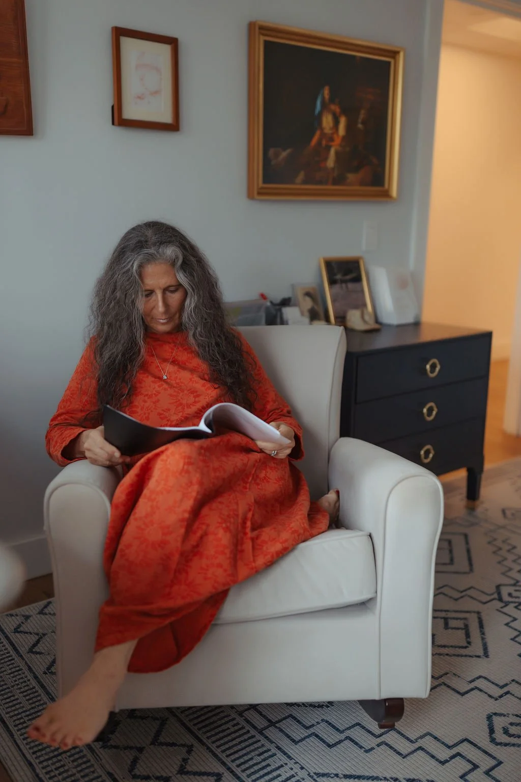 A woman with long curly gray hair, wearing an orange dress, sitting on a white armchair and reading a book in a living room. There are framed pictures and paintings on the wall and a black dresser with decorative items behind her.