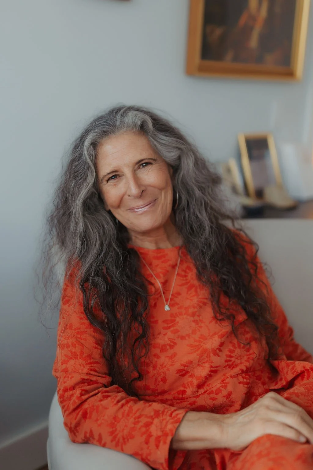 A smiling woman with long, curly gray hair, wearing an orange patterned top with a necklace, sitting indoors with framed pictures on the wall behind her.