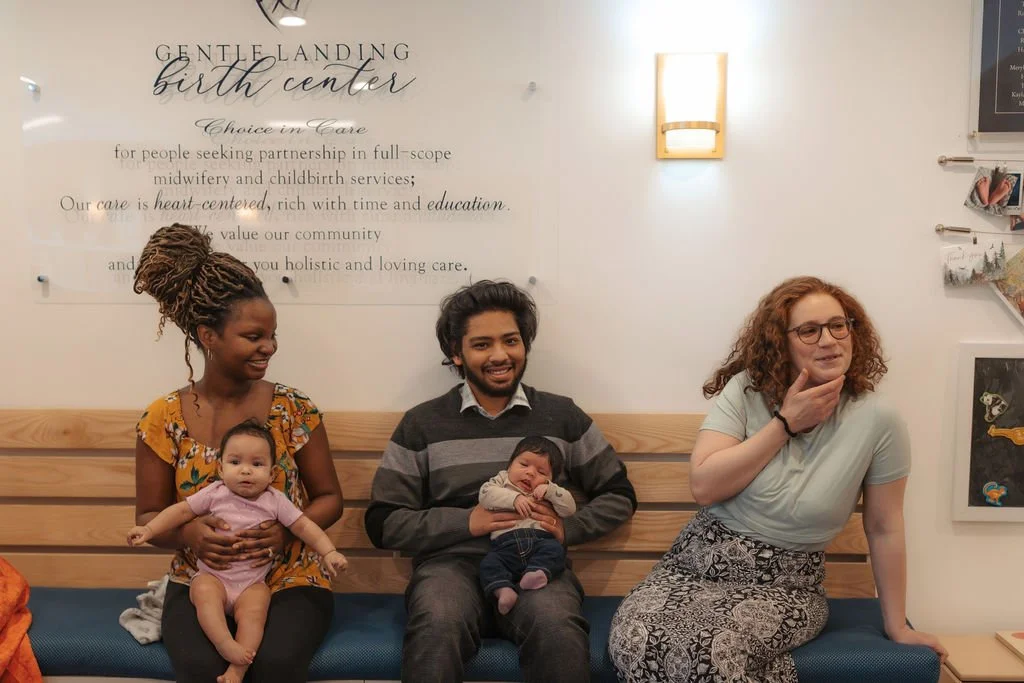 A group of four people sitting on a bench in a birth center. On the left, a woman with braided hair is holding a baby girl. Next to her, a man with dark hair and a beard is holding a sleeping infant. On the right, a woman with red curly hair and glas