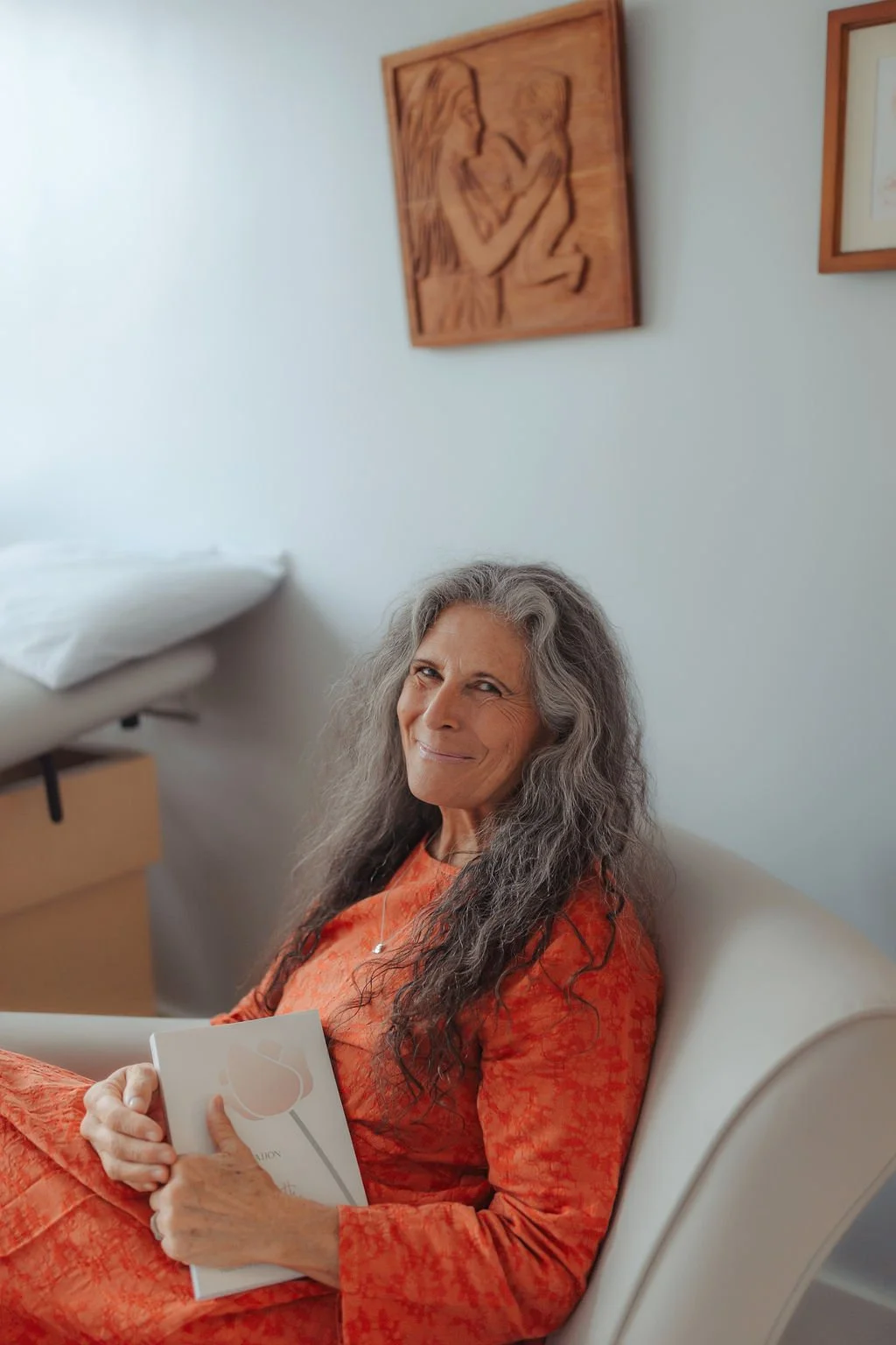 An elderly woman with long gray hair is sitting on a hospital bed, holding a booklet, wearing an orange dress, and smiling at the camera. The room has light-colored walls with framed art, and there is a pillow and a bedside table nearby.