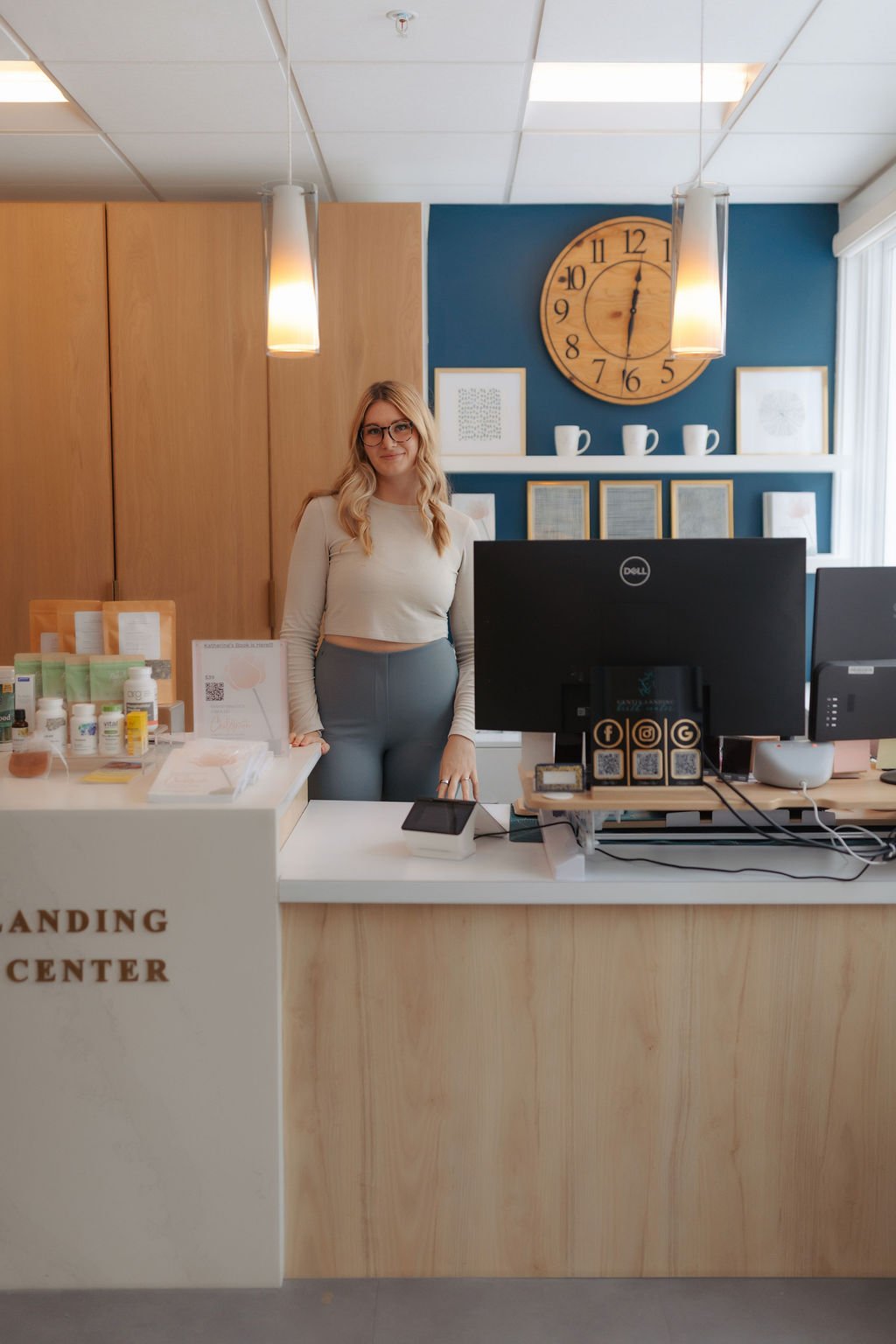 A young woman with blonde hair, glasses, wearing a light-colored long sleeve shirt and gray pants, standing behind a reception desk in an office or wellness center with pills and bottles on the counter. The background features a blue wall with a larg