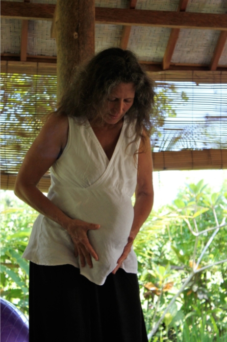A woman with curly hair wearing a sleeveless white top and black pants, standing outdoors in a tropical setting, holding her stomach.