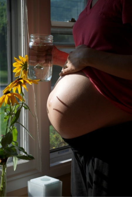 A pregnant person standing by a window holding a glass of water with flowers nearby, sunlight illuminating their belly.