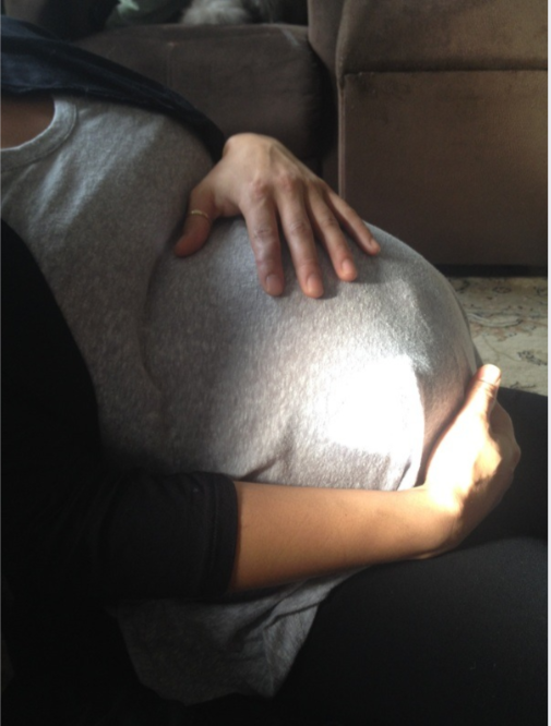 A pregnant woman with a hand resting on her belly and a child's arm and hand embracing her belly.