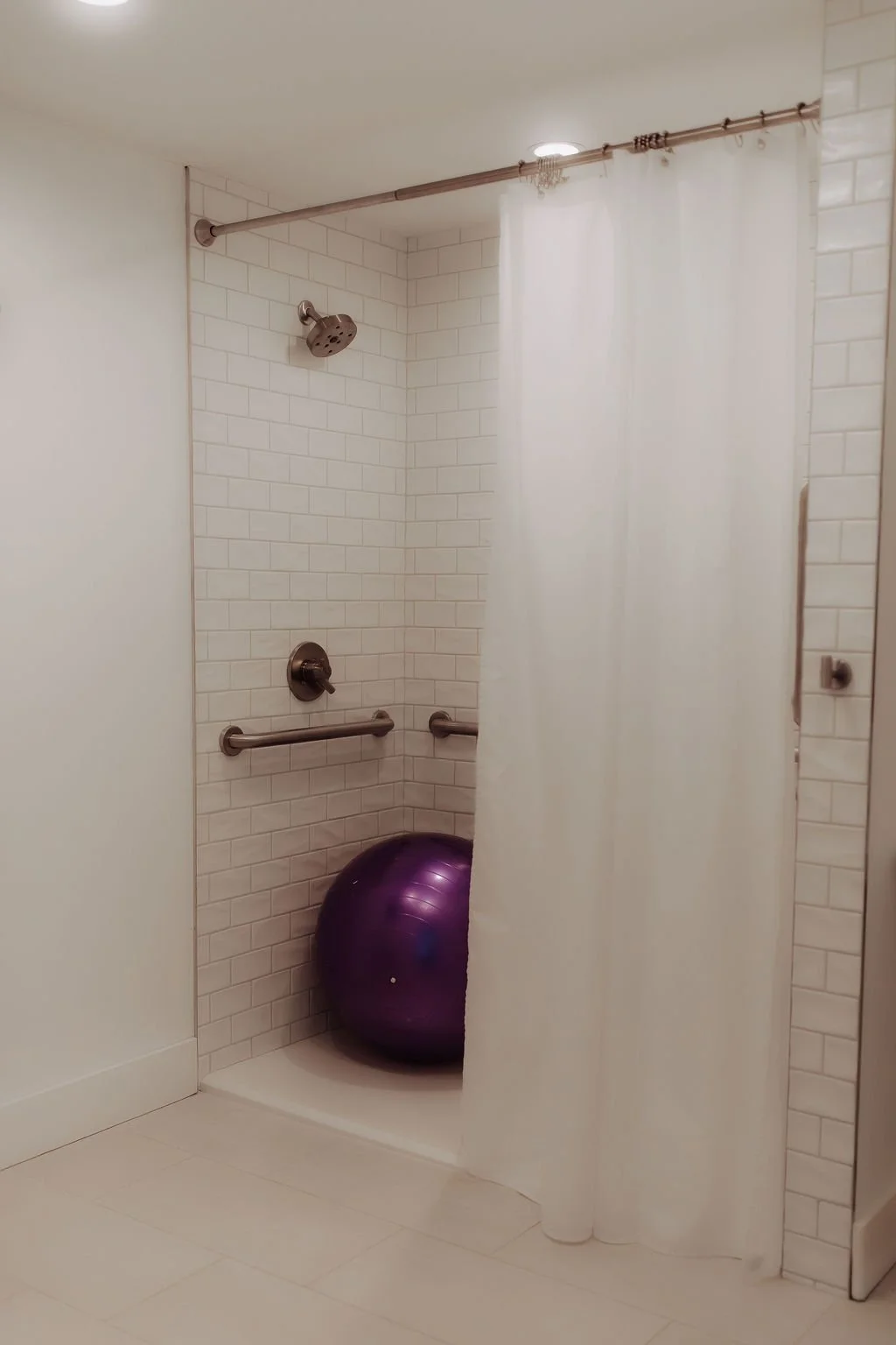 Walk-in shower with white subway tiles, a purple exercise ball, metal grab bars, a white shower curtain, and a partially visible wall-mounted mirror.