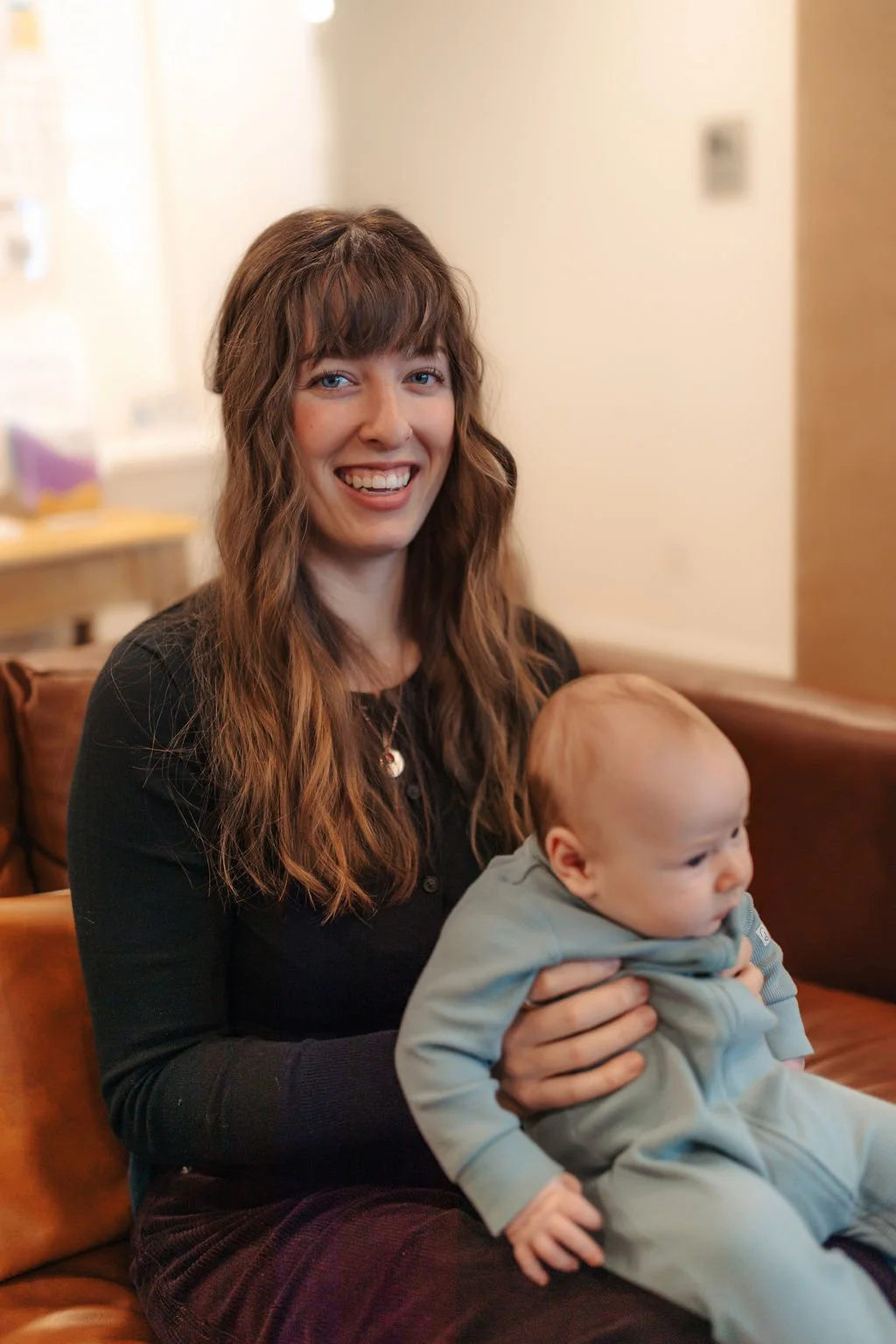 A woman with long wavy brown hair sitting on a brown leather couch, smiling, while holding a young child with a bald head wearing a light blue outfit.