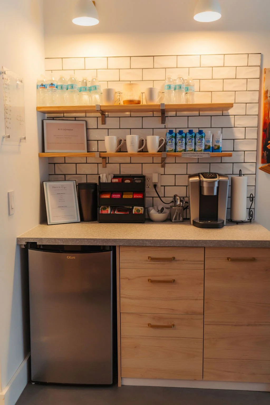 Coffee station with shelves holding water bottles, cups, juice cartons, and a framed certificate, with a coffee maker, paper towel roll, and small refrigerator below on a beige countertop.