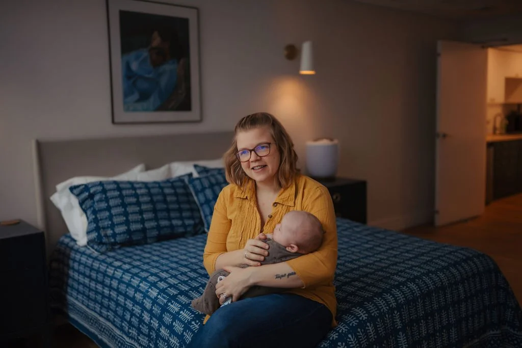 Woman with glasses in a yellow shirt holding a sleeping baby on her lap in a bedroom.