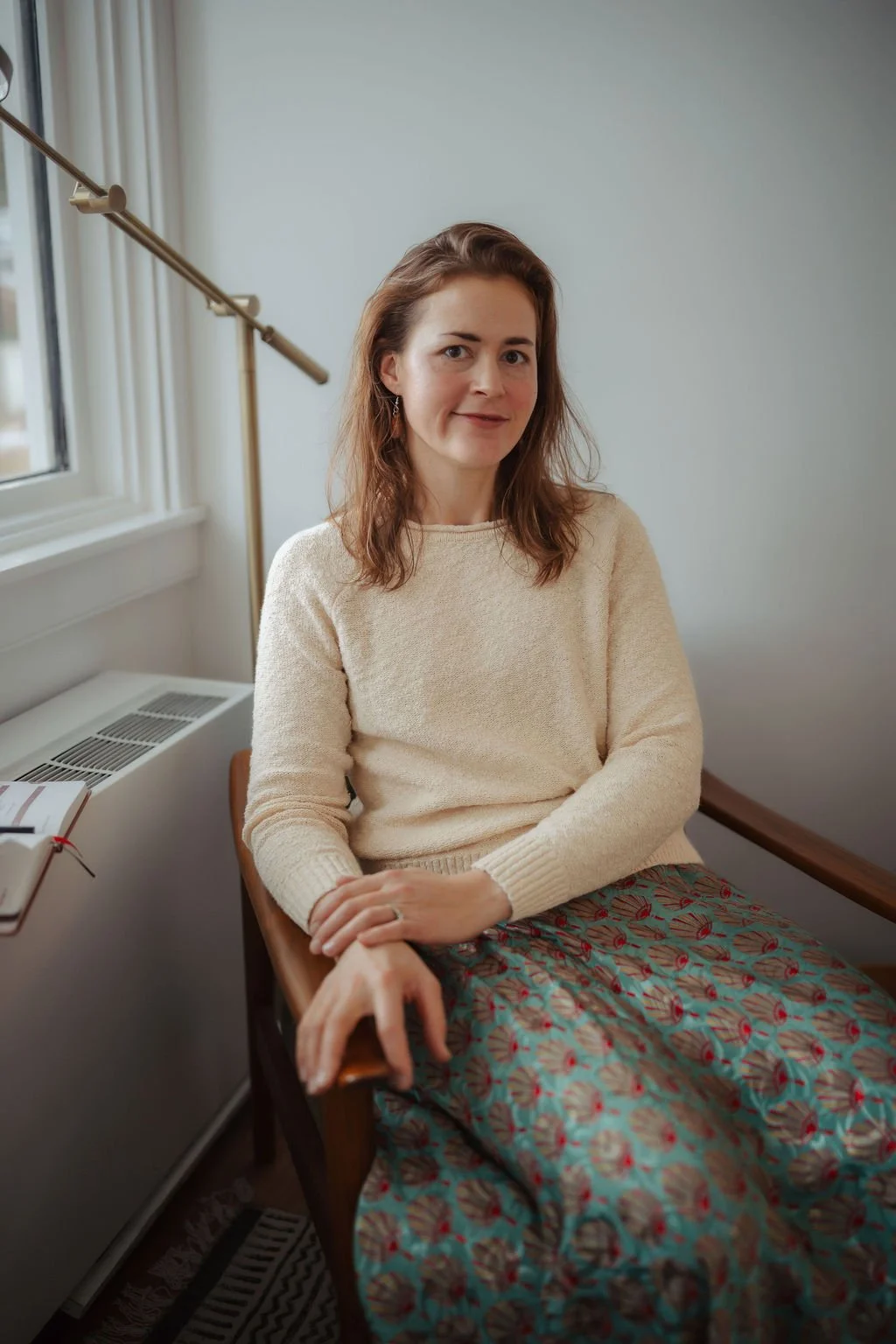 A woman with brown hair wearing a cream sweater and patterned skirt sitting in a wooden chair near a window in a cozy room.