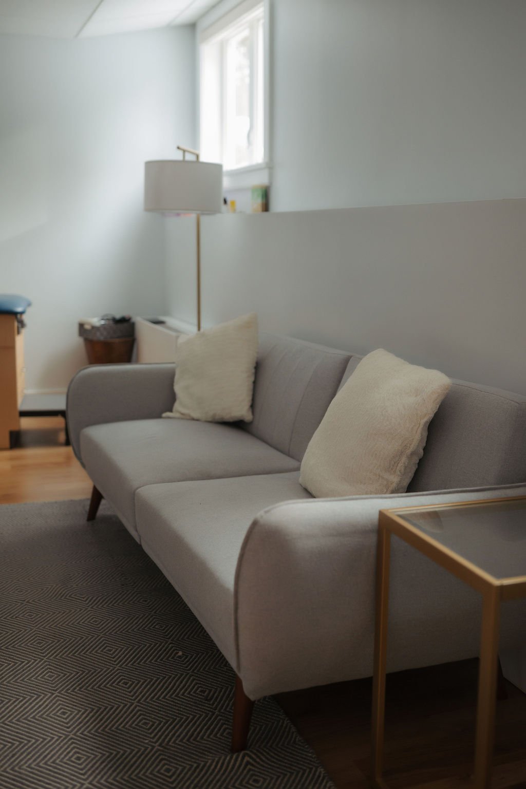 Living room with a light gray sofa, two beige pillows, a small wooden side table with a glass top, a floor lamp behind the sofa, and a window above the lamp providing natural light.