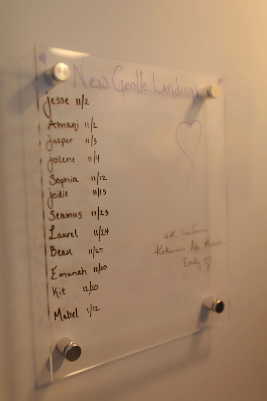 A clear acrylic board with handwritten names and dates, titled 'New Gentle Handings,' attached to a wall with four metallic bolts, and a purple heart drawing on the right side.