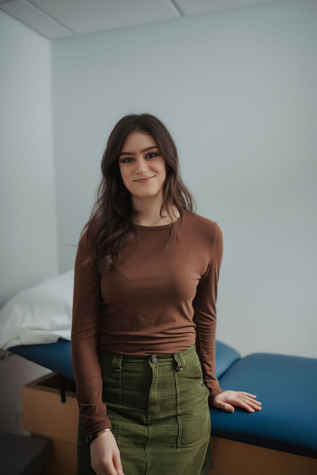 A young woman with long, dark brown hair and light skin standing in a room with white walls and a bed in the background, smiling at the camera. She is wearing a brown long-sleeve top and an olive green skirt.