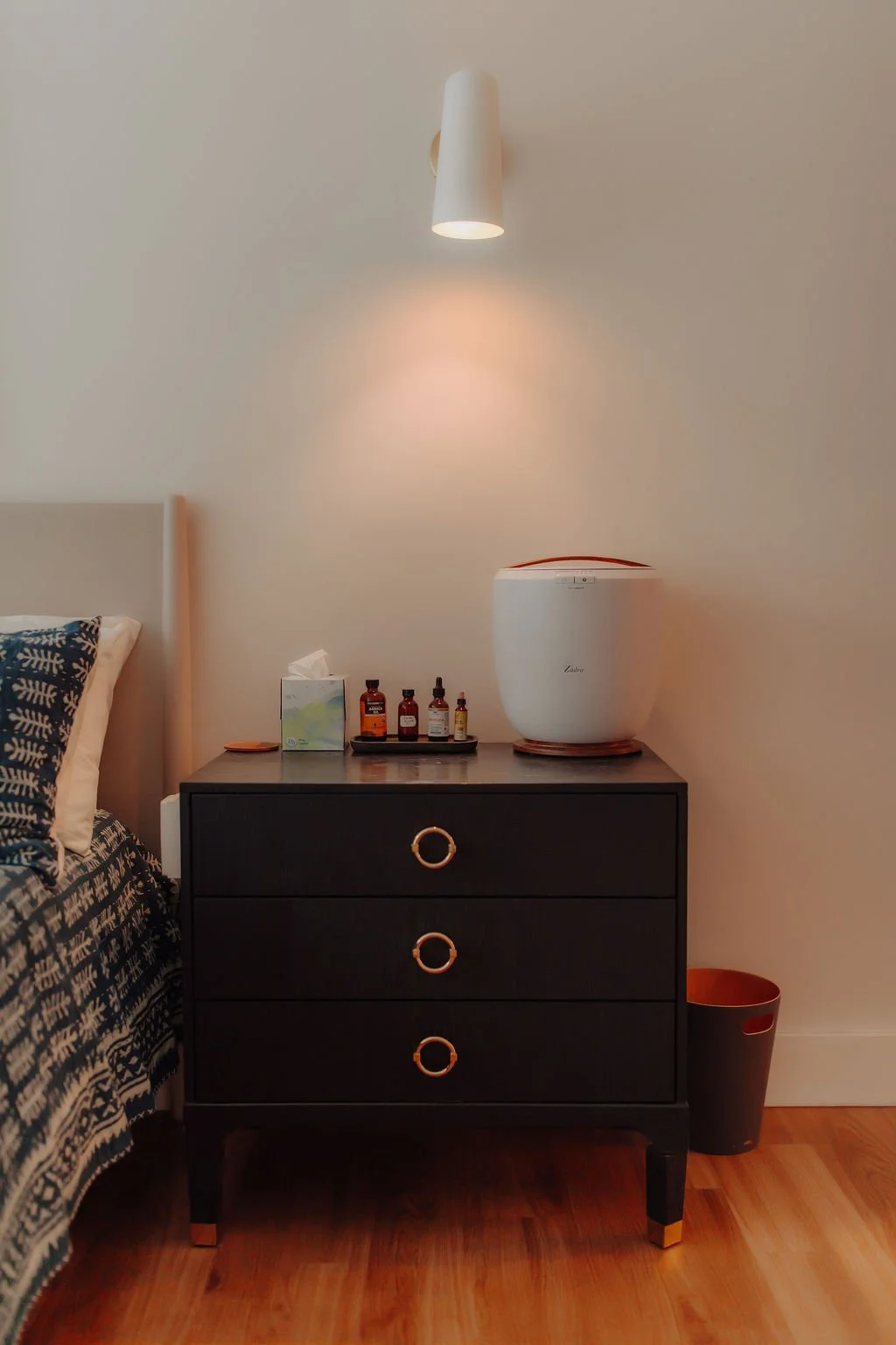 A black bedside table with three drawers featuring gold ring handles, on a hardwood floor. On top of the table are essential oils, tissue box, and a white air purifier. Above, a modern white wall sconce illuminates the wall.