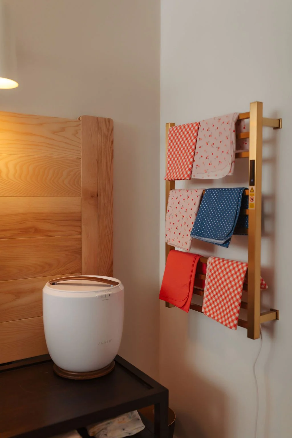 A wooden headboard of a bed, a white humidifier or air purifier on a black table, and a wooden towel rack with colorful cloths hanging on a white wall.