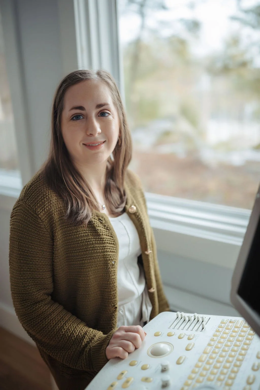 A young woman smiling at the camera inside a building, with an ultrasound machine in front of her.