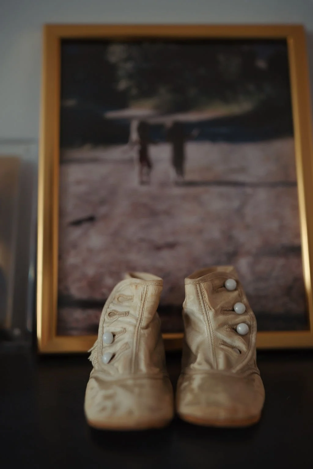 A pair of vintage cream-colored baby shoes with white buttons, placed on a flat surface. Behind them, slightly out of focus, a framed photo depicts two children walking outdoors near a body of water with trees in the background.