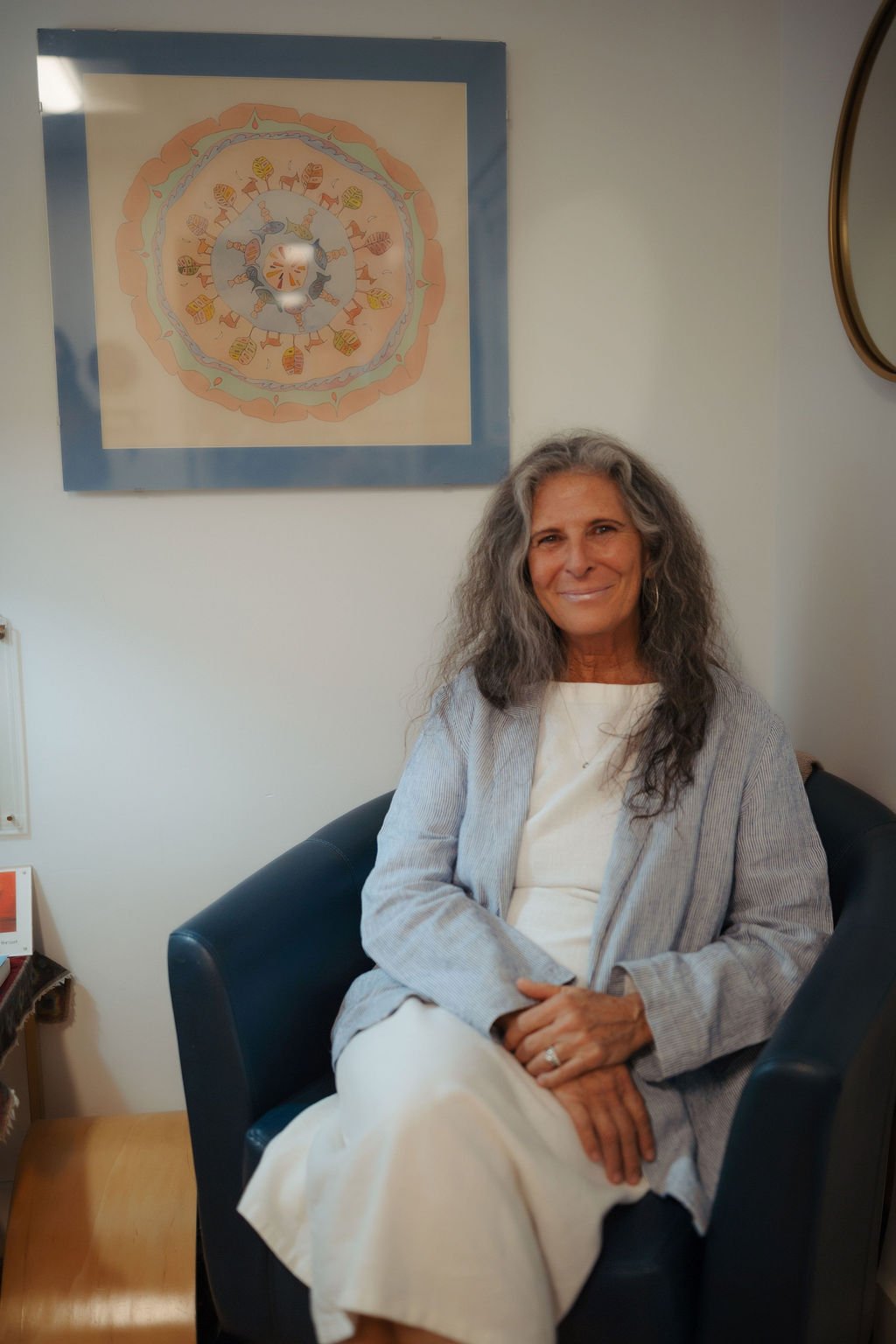 A woman with long, wavy gray hair smiling and sitting on a dark blue armchair in a waiting room. She is wearing a light-colored dress and a gray striped blazer. Behind her is a wall with a circular artwork featuring colorful, whimsical animal and nat