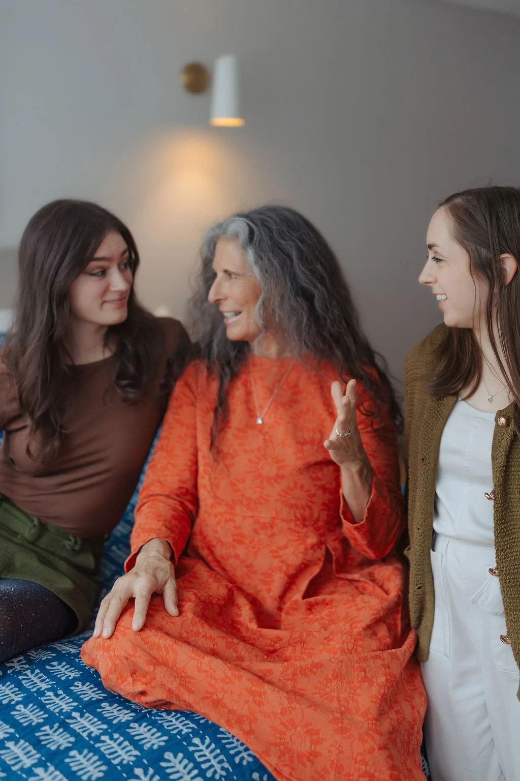 A woman with gray hair wearing an orange dress is talking and smiling while sitting on a bed, flanked by two younger women, one with brown hair and the other with light brown hair, all engaged in a conversation.