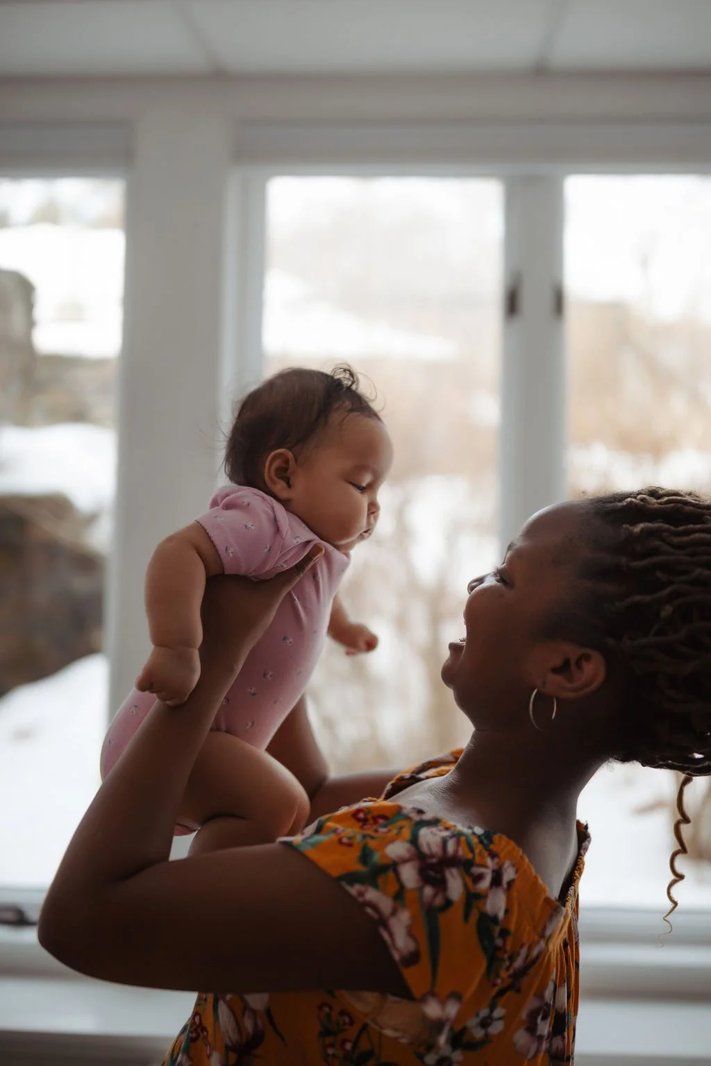 A woman lifting a baby girl inside a room with large windows, snow visible outside.