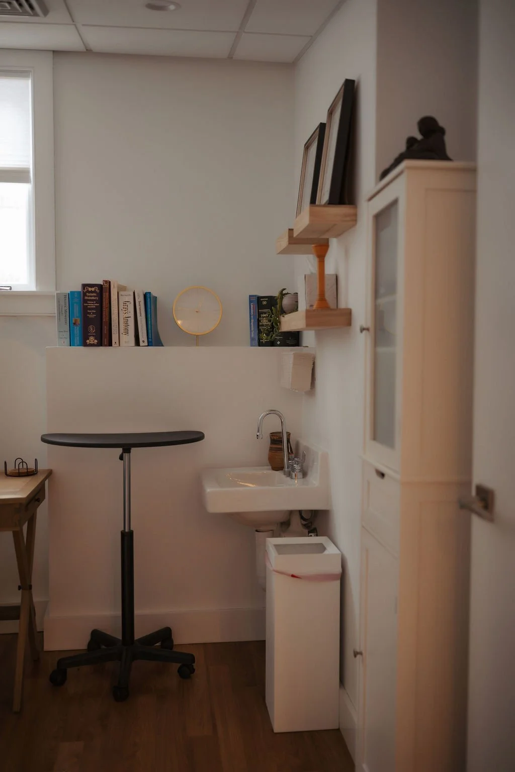 A small corner of a room with a window, a black rolling stool, a small wooden table, a wall-mounted sink with a brown soap dispenser, and a white cabinet. Floating shelves hold books, framed pictures, a gold clock, and decorative items.