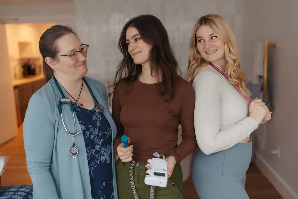Three women, including a healthcare professional with a stethoscope, standing together and smiling inside a medical facility, possibly a clinic or hospital.