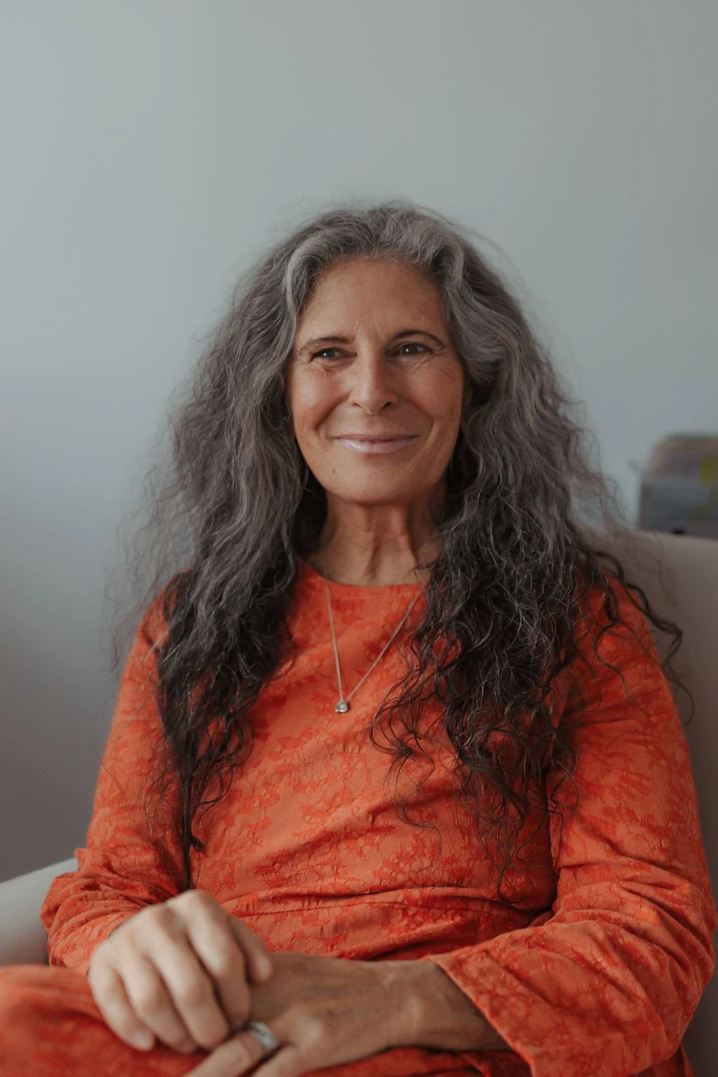 A smiling mature woman with long curly gray hair, wearing an orange tie-dye top and a silver necklace, sitting indoors against a plain light-colored wall.