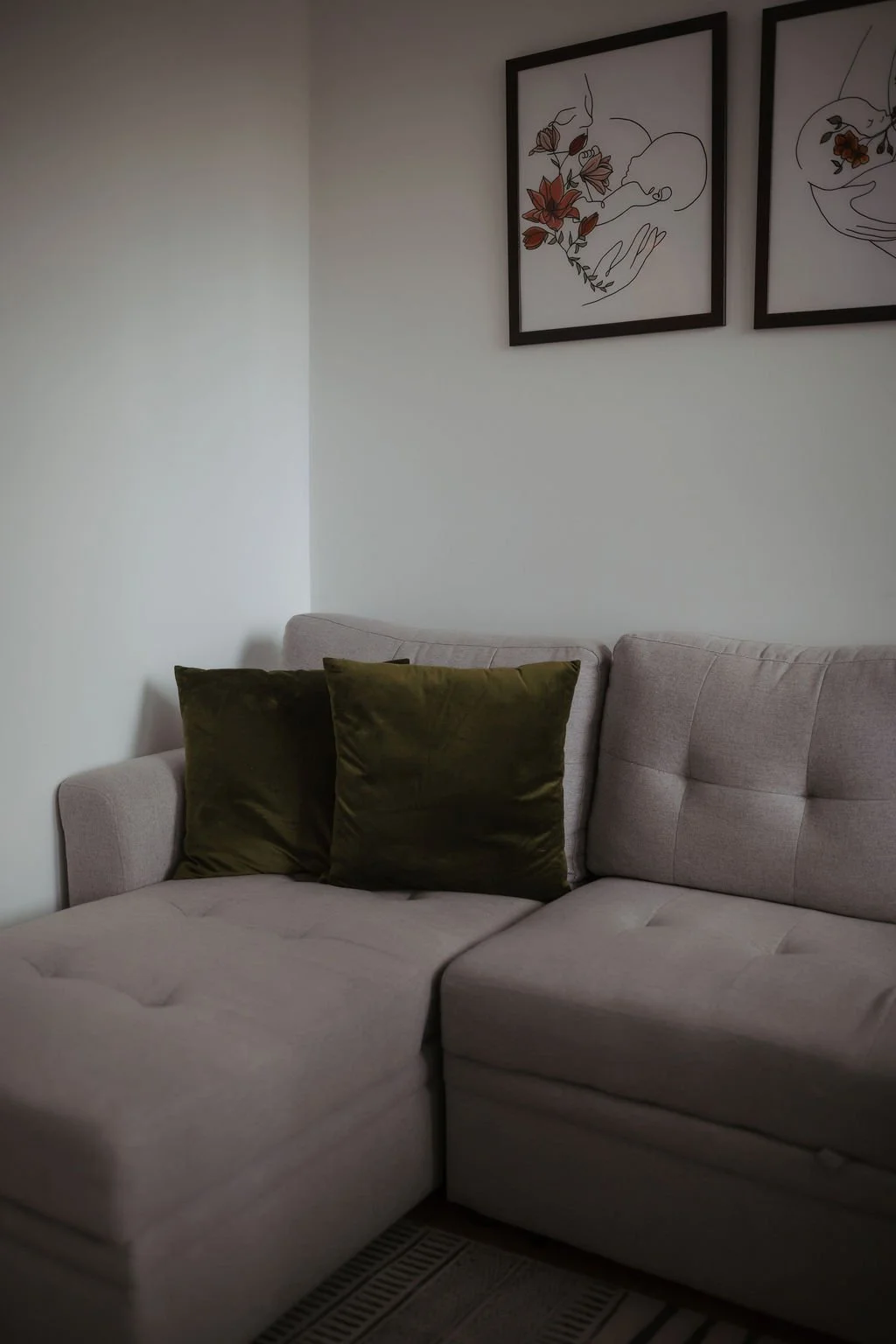 A beige sectional sofa with two dark green pillows, a white wall with two framed floral line art drawings, and a patterned rug covering part of the floor.